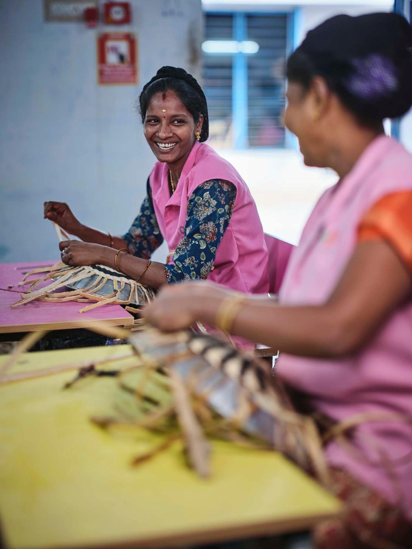 Two people smile as they weave baskets side by side in an indoor setting.