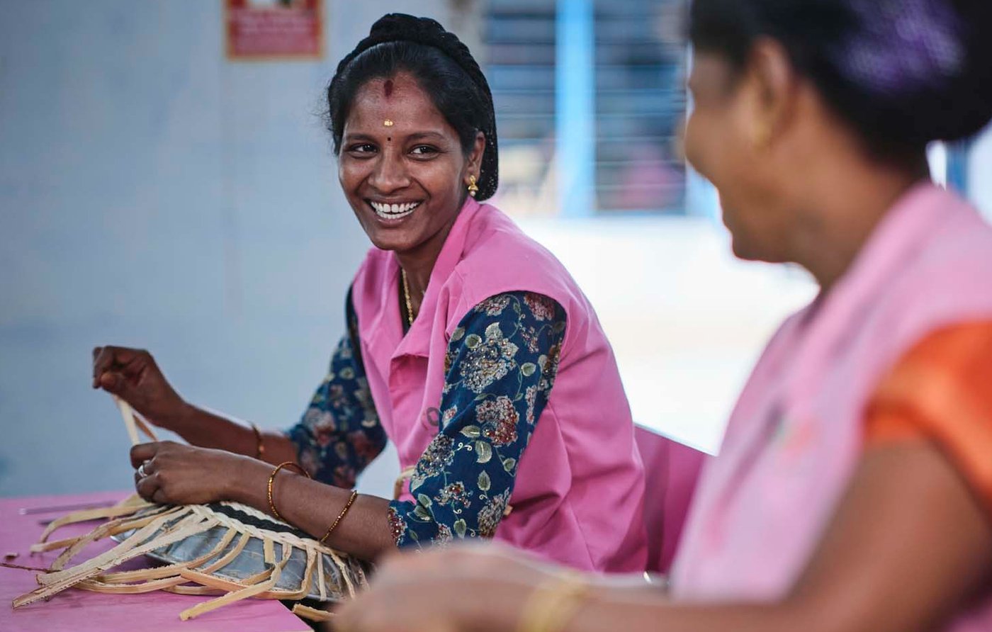 Two people smile as they weave baskets side by side in an indoor setting.