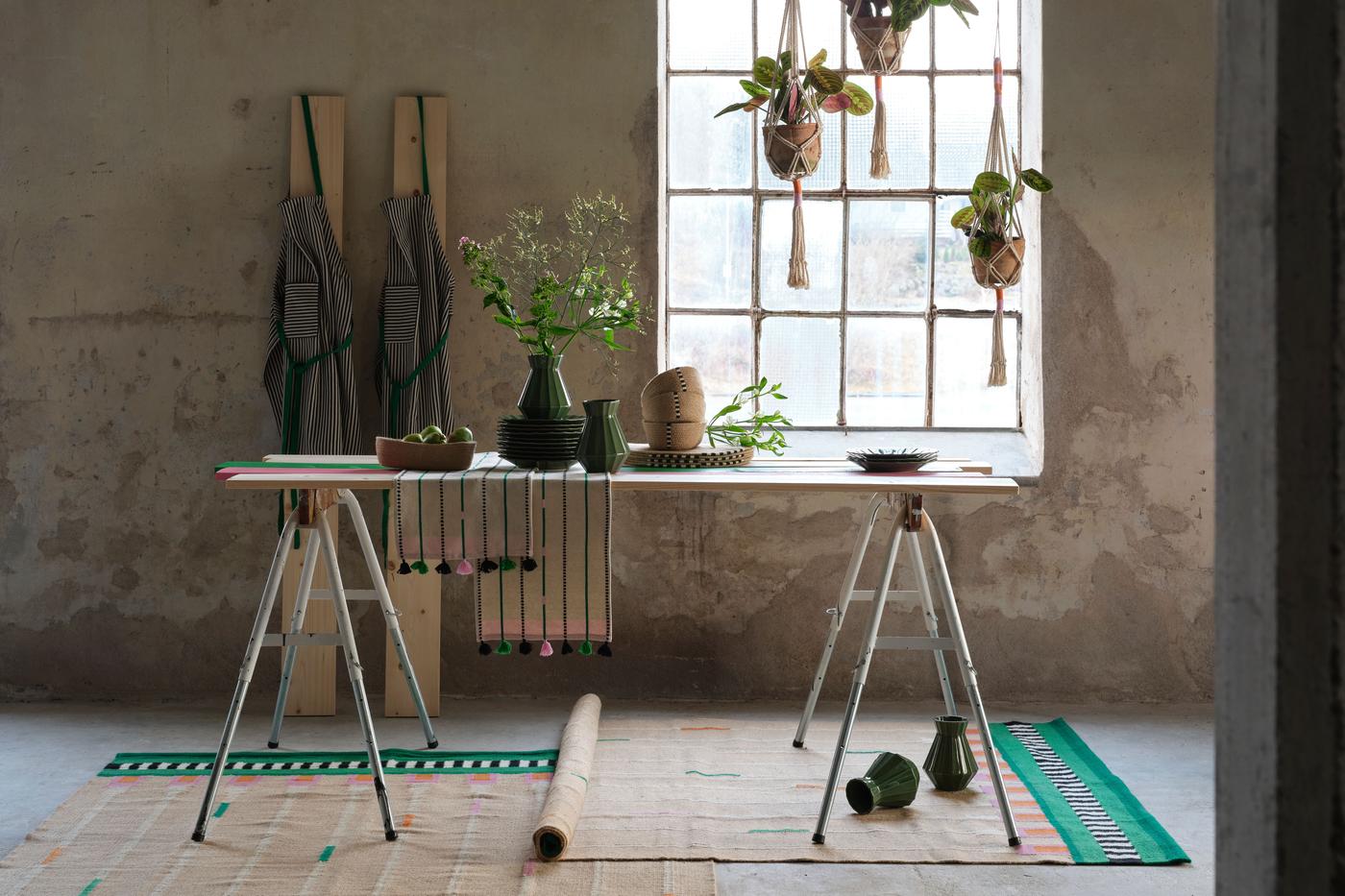 Vases, aprons, rugs and hanging plant holders from the MÄVINN collection are shown in a room setting, displayed on and around a table in front of a window..