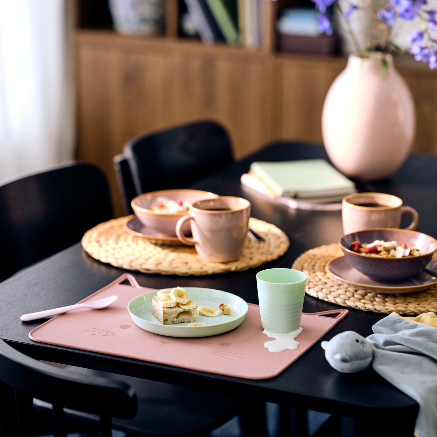 A breakfast table set with a pink children's cat shaped placemat, mint green tableware and pink dinnerware set in the back on natural placemats.