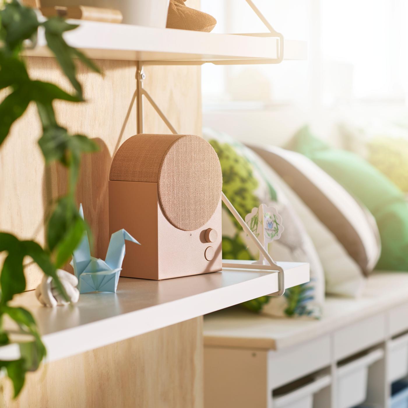 A close-up of a pink speaker on a light wooden shelf, with greenery and small decorations nearby.