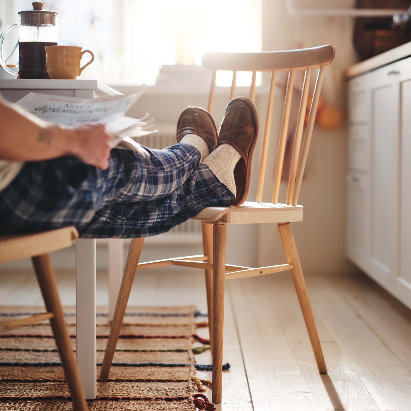 A person sits in a light, wood kitchen in their pyjamas with their feet on a wooden chair.