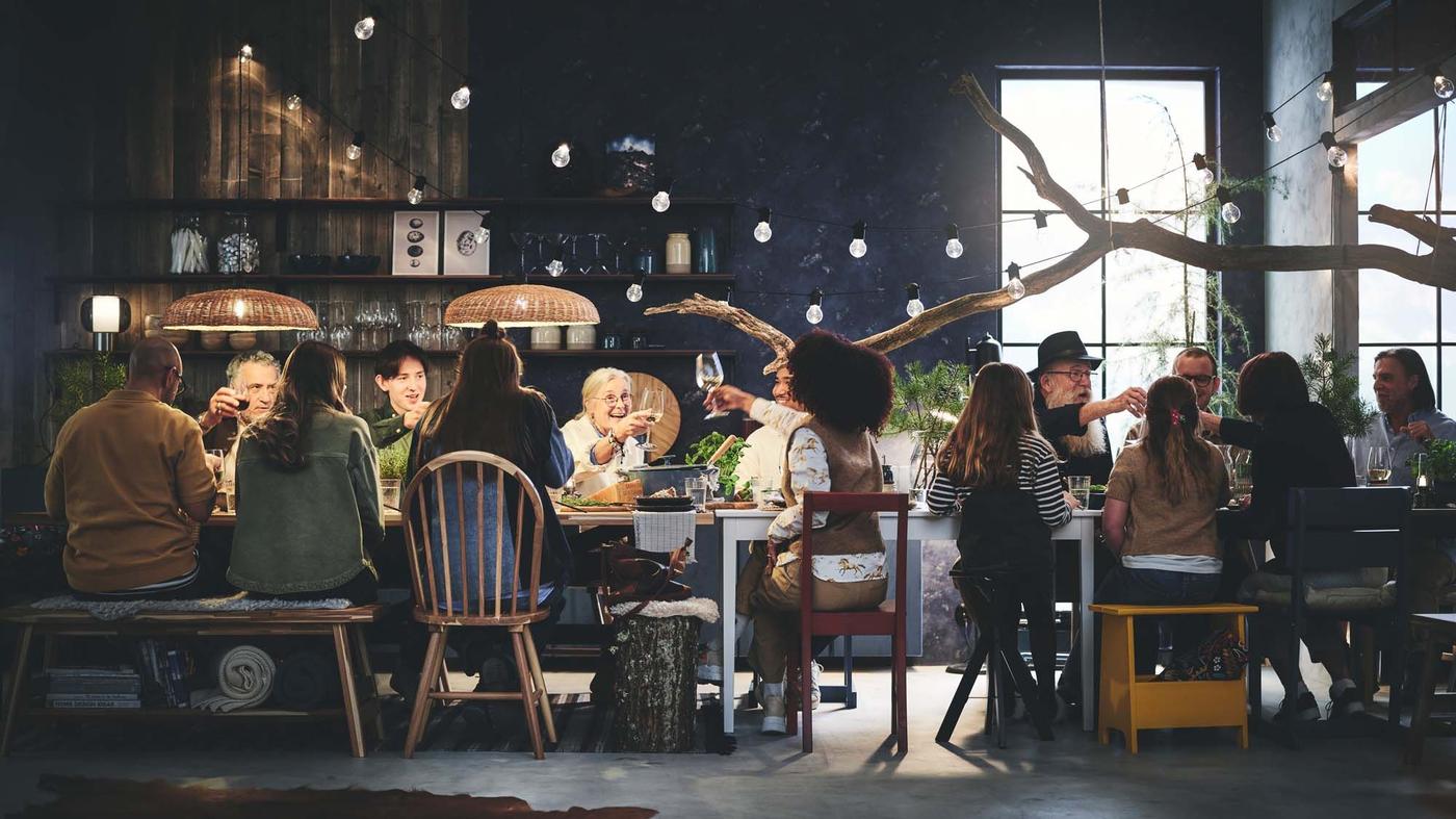 A large group of people sit together at a long table in a dark coloured dining area. The atmosphere is jolly and celebratory as they eat and drink together.