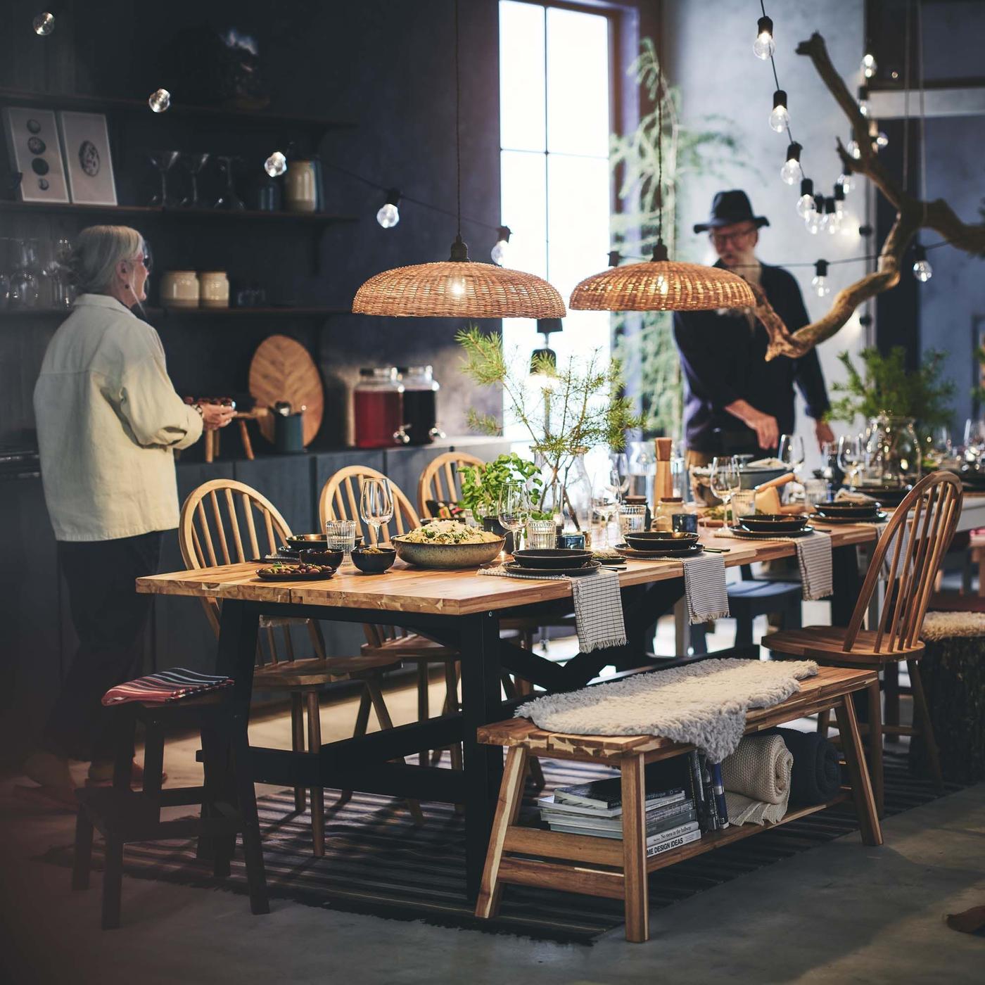 Two people add plates of food to a long, rustic dining table lined with many chairs.