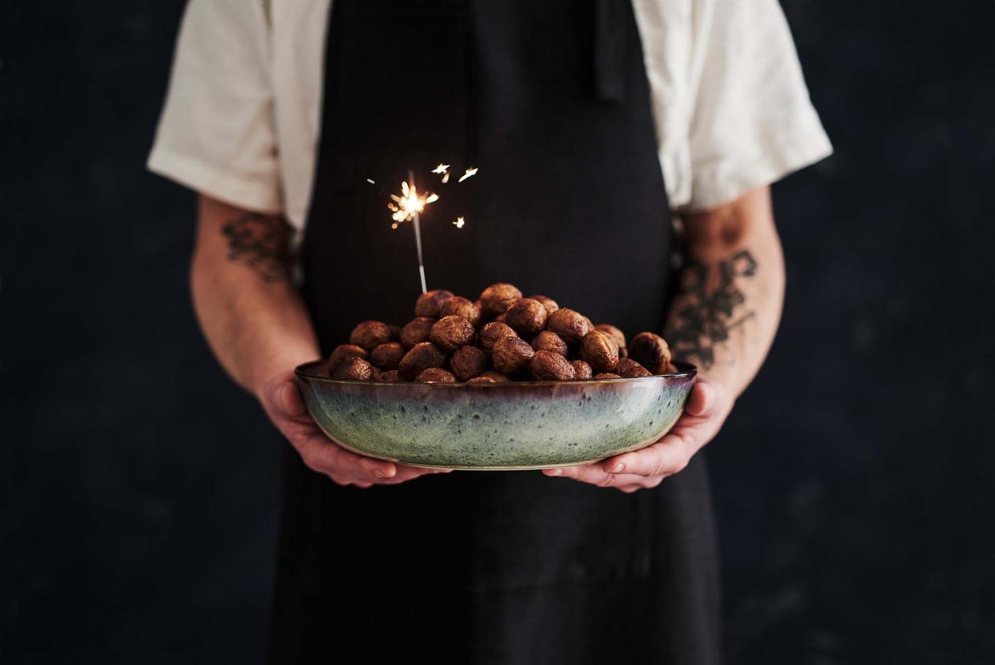 A person wearing an apron holds a plate of meatballs with an illuminated sparkler, adding a festive look to the dish.