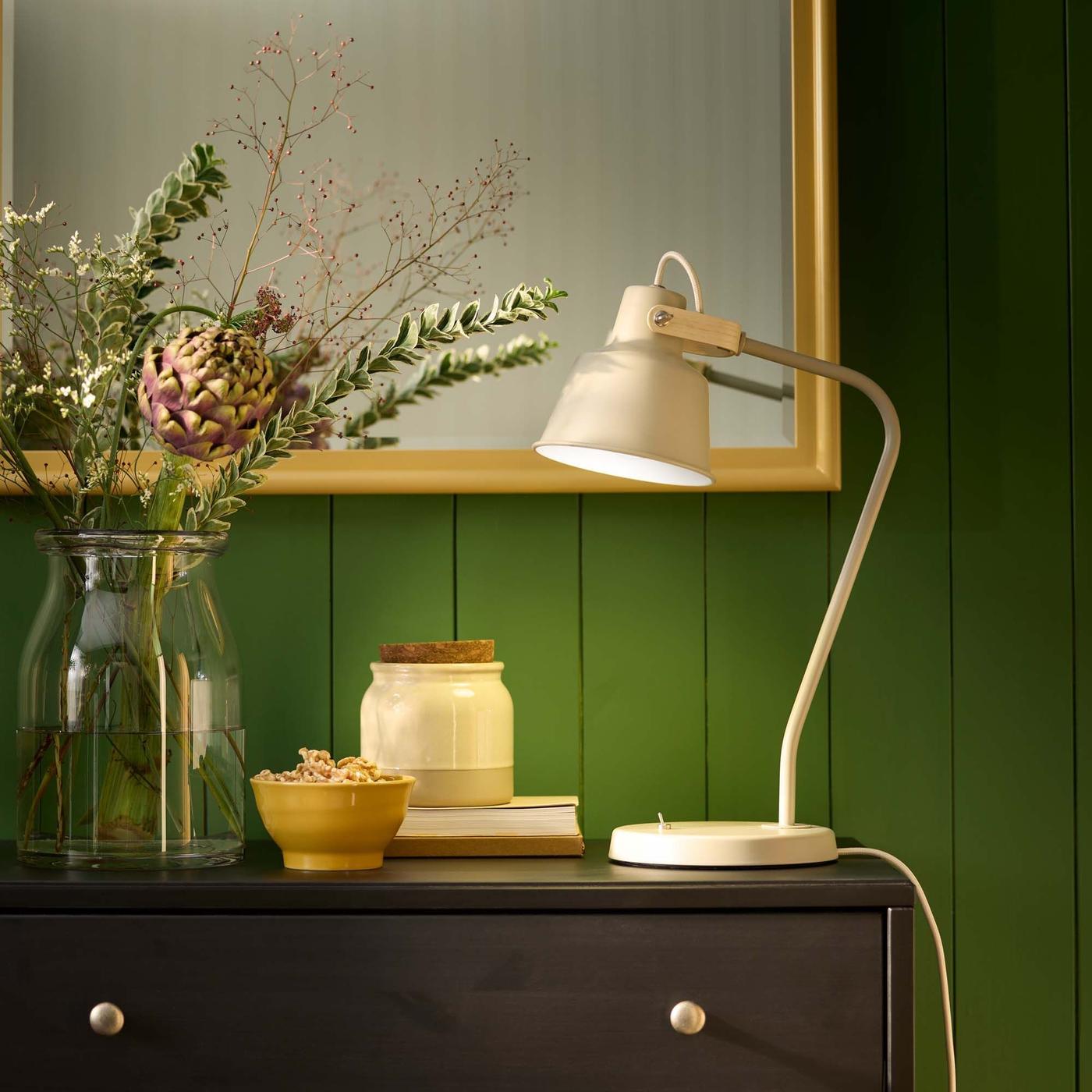 A cream SKÖMAKT reading lamp next to a jar and a vase of flowers illuminates a chest of drawers.