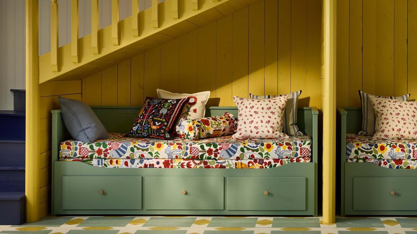A green HEMNES daybed under a yellow staircase, propped with colourful patterned pillows and bedlinen.
