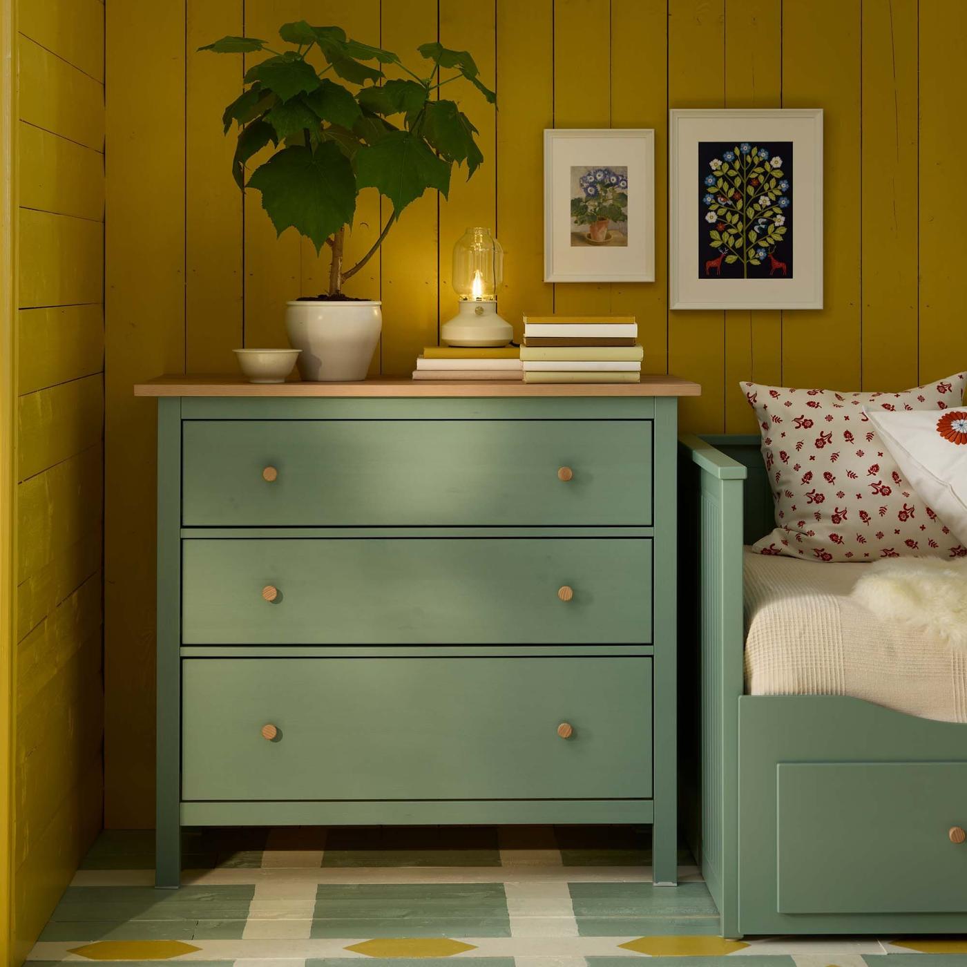 A green HEMNES chest of drawers in a corner, with a plant, a lamp and books on top, against a rustic yellow wall.