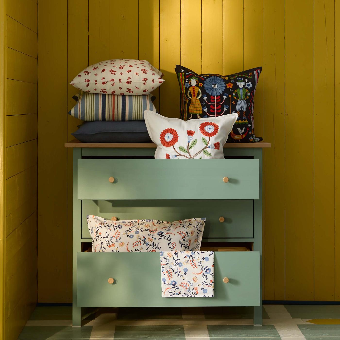A green chest of drawers with cushions inside drawers and on top, against a yellow wood background.