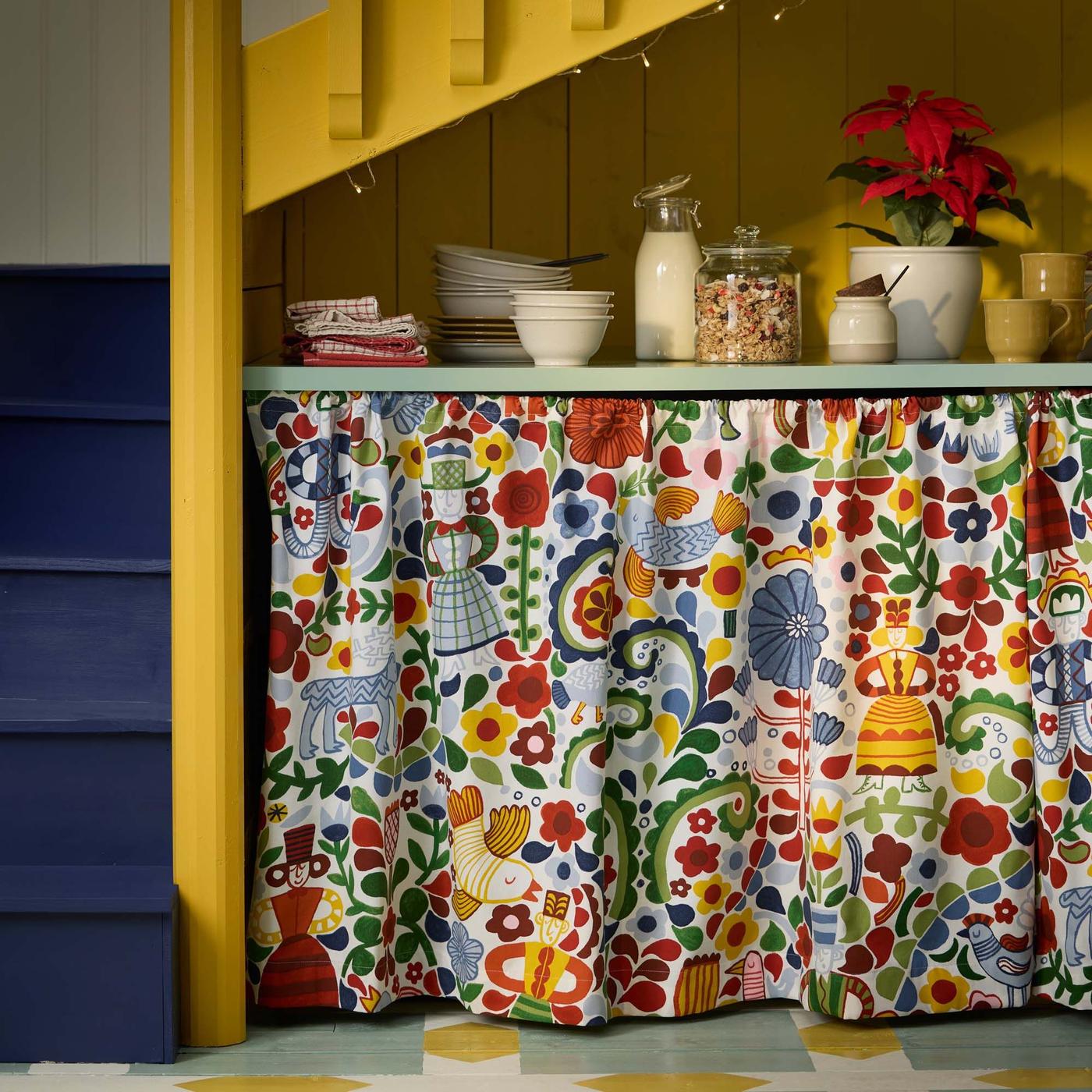A small breakfast serving area under yellow and blue stairs. A colourful curtain conceals shelves below.
