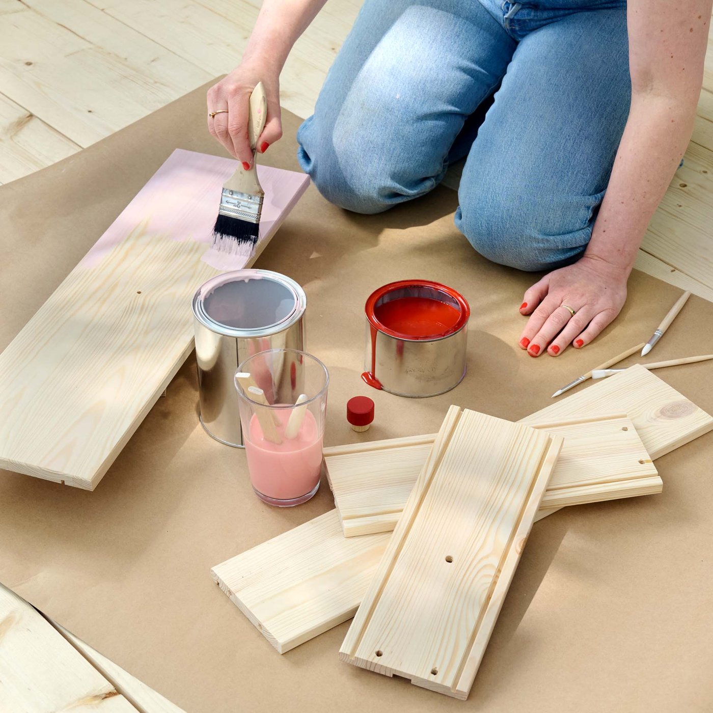 A person in light blue jeans is kneeling on the floor painting a light wood panel pale pink.