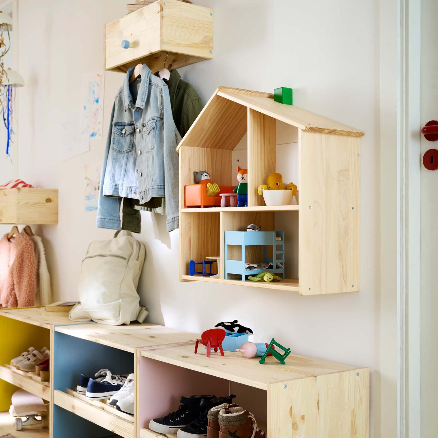 The wall of a hallway with a painted RAGGBERG bench with shoe storage below, a RAGGBERG coat rack with jackets above and a FLISAT doll's house filled with furniture.