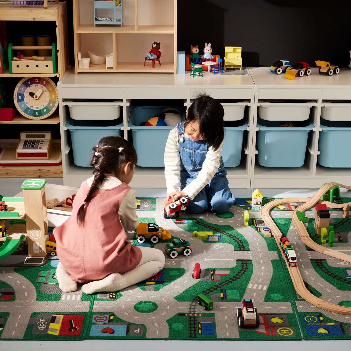 Two children sit on a playmat designed like roads in a bedroom. They are playing with cars on the rug.
