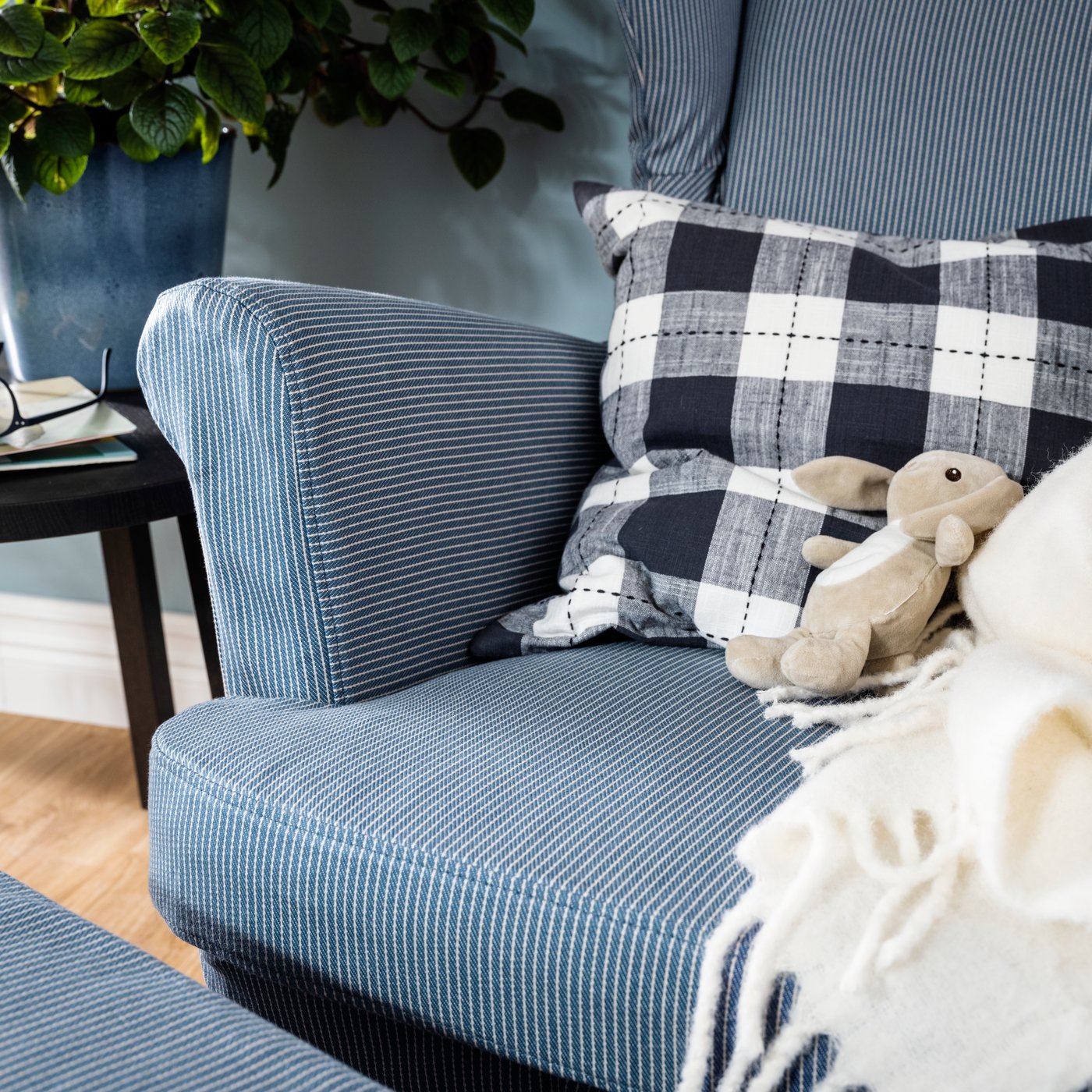 A close-up of a checked cushion, soft toy rabbit and white blanket on a blue and white striped sofa.