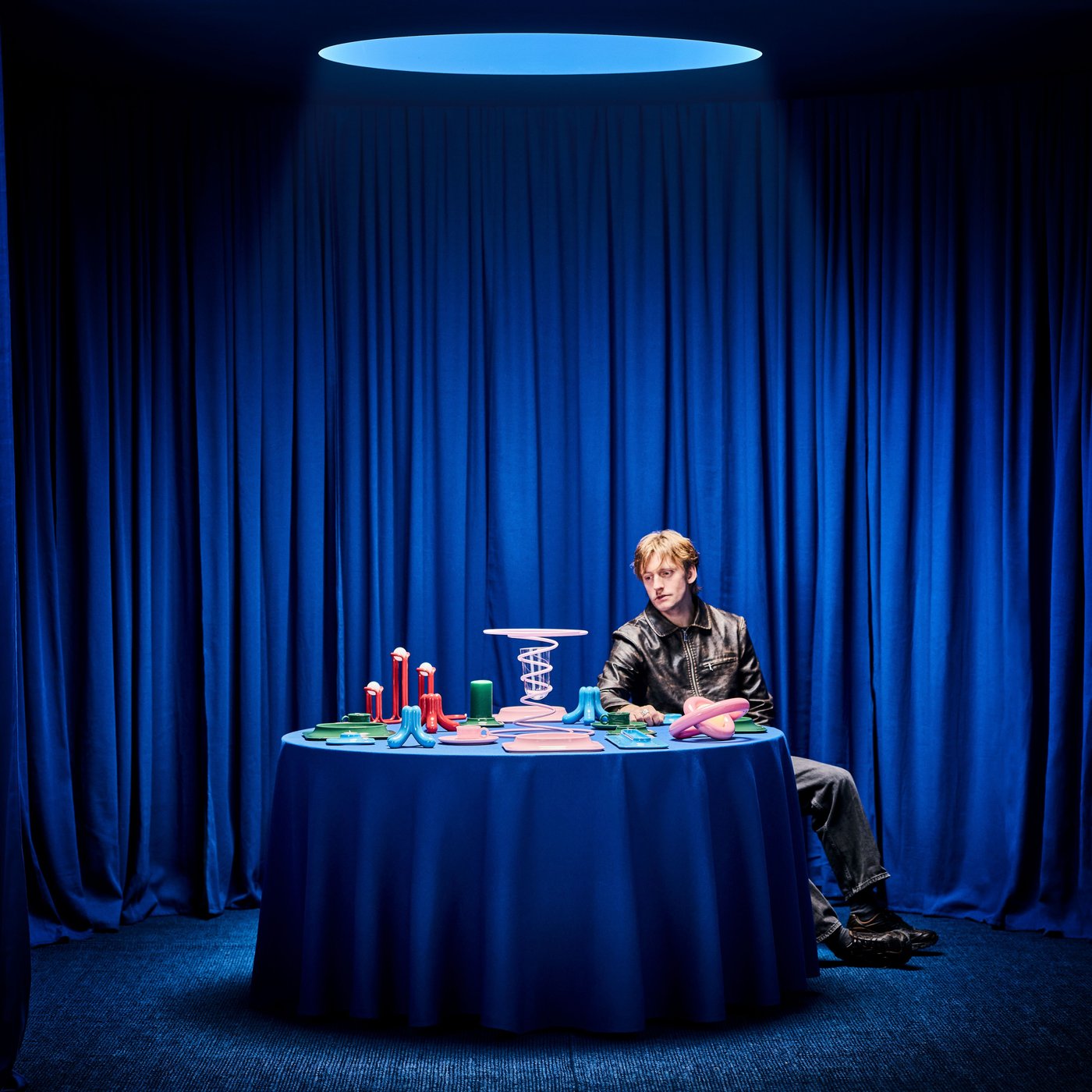 Gustaf Westman seated by a blue-clothed table of multicolored collection products, glancing down, with blue curtains behind.