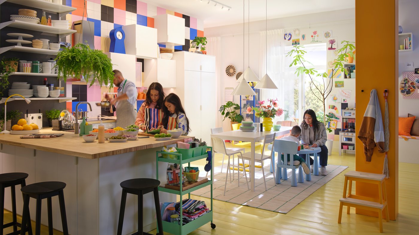 A large colourful family kitchen. Two children stand at a kitchen island baking, while dad puts something in the oven. At a small table a mother and young child are drawing.