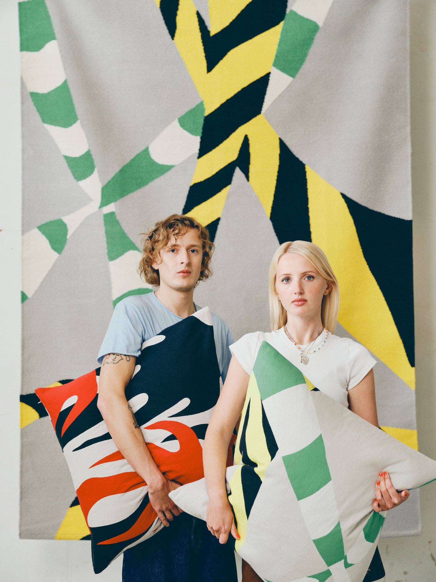 Two people each holding a large pillow in front of them with abstract design, set against the backdrop of a large rug hung on the wall in abstract design.