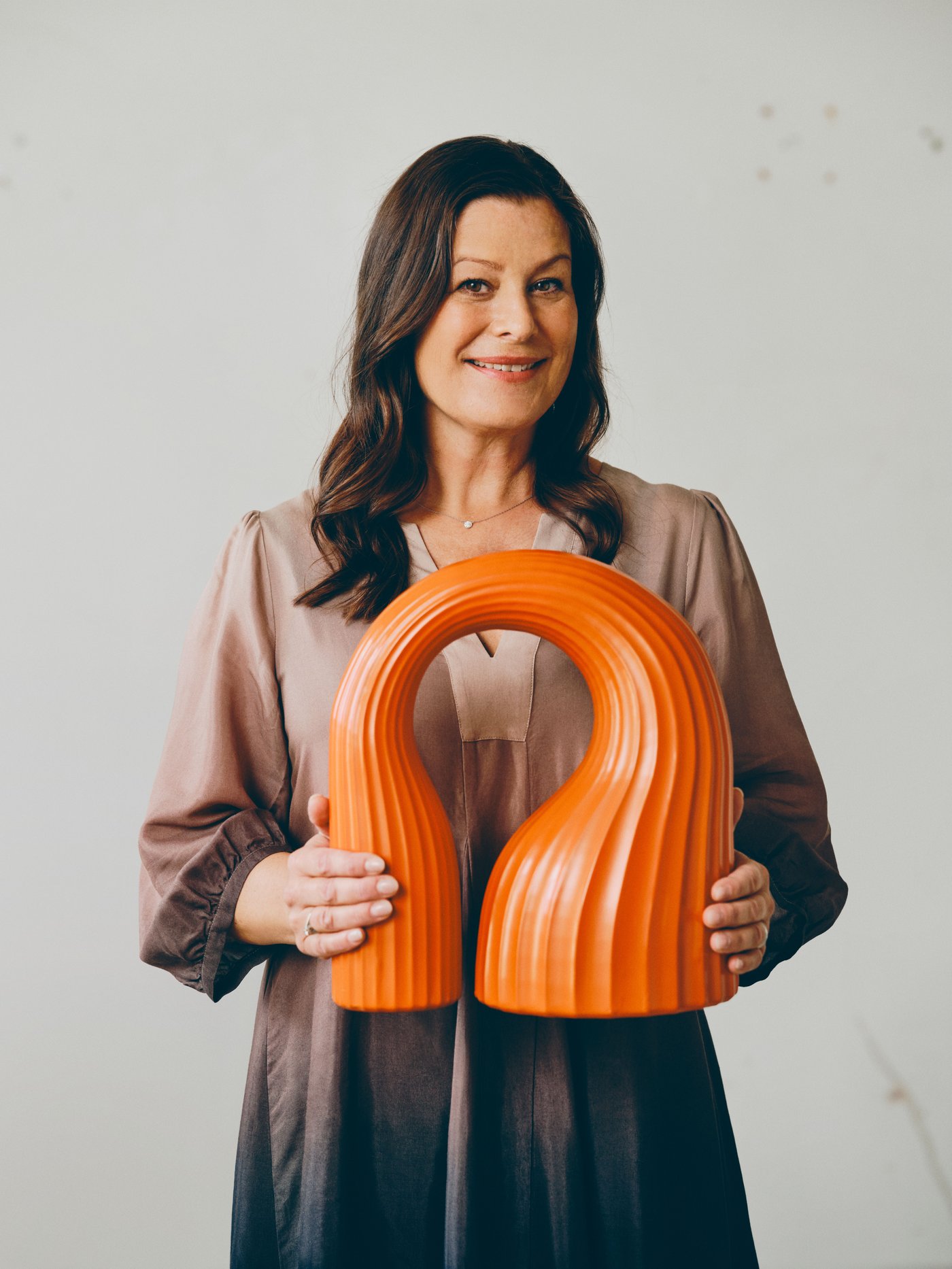 Woman standing and holding an orange sculpture with two hands against a light backdrop.