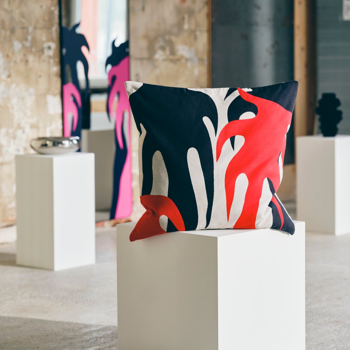 Large pillow with abstract design displayed on a white plinth in an industrial gallery, with a mirror leaning against the wall in the background.