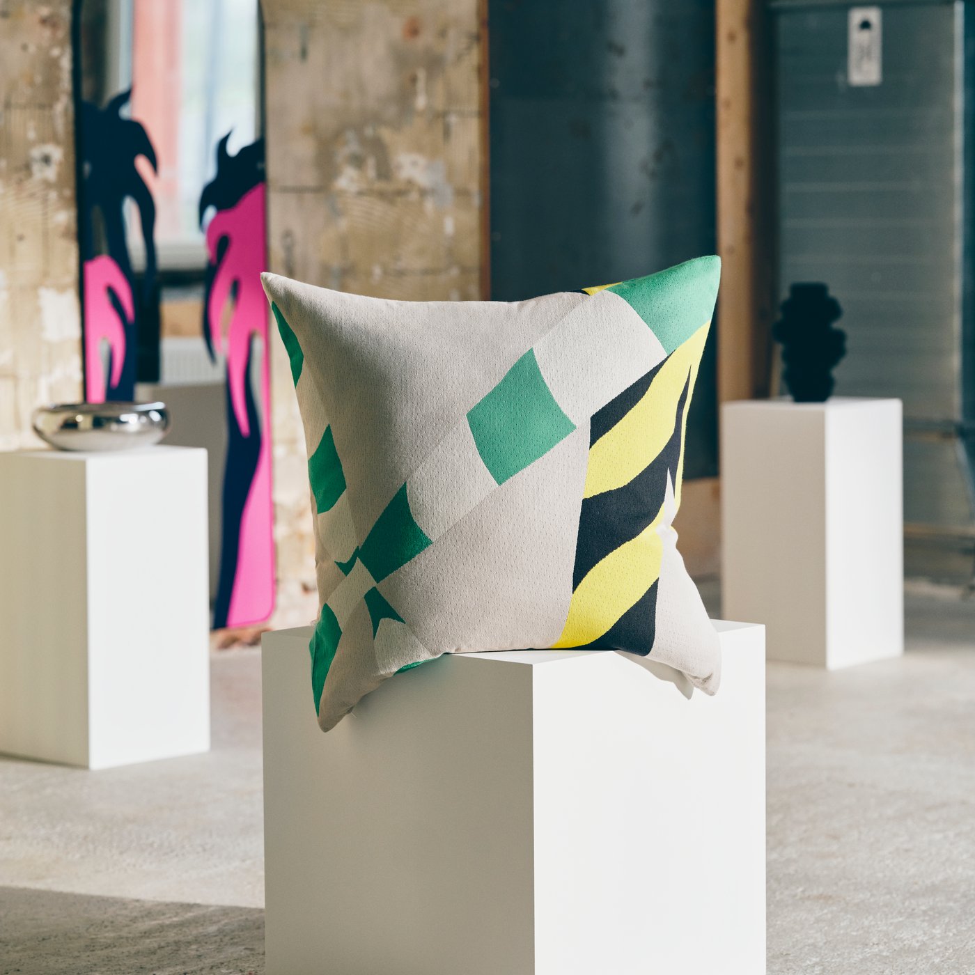Large pillow with abstract design displayed on a white plinth in an industrial gallery, with a mirror leaning against the wall in the background.. 