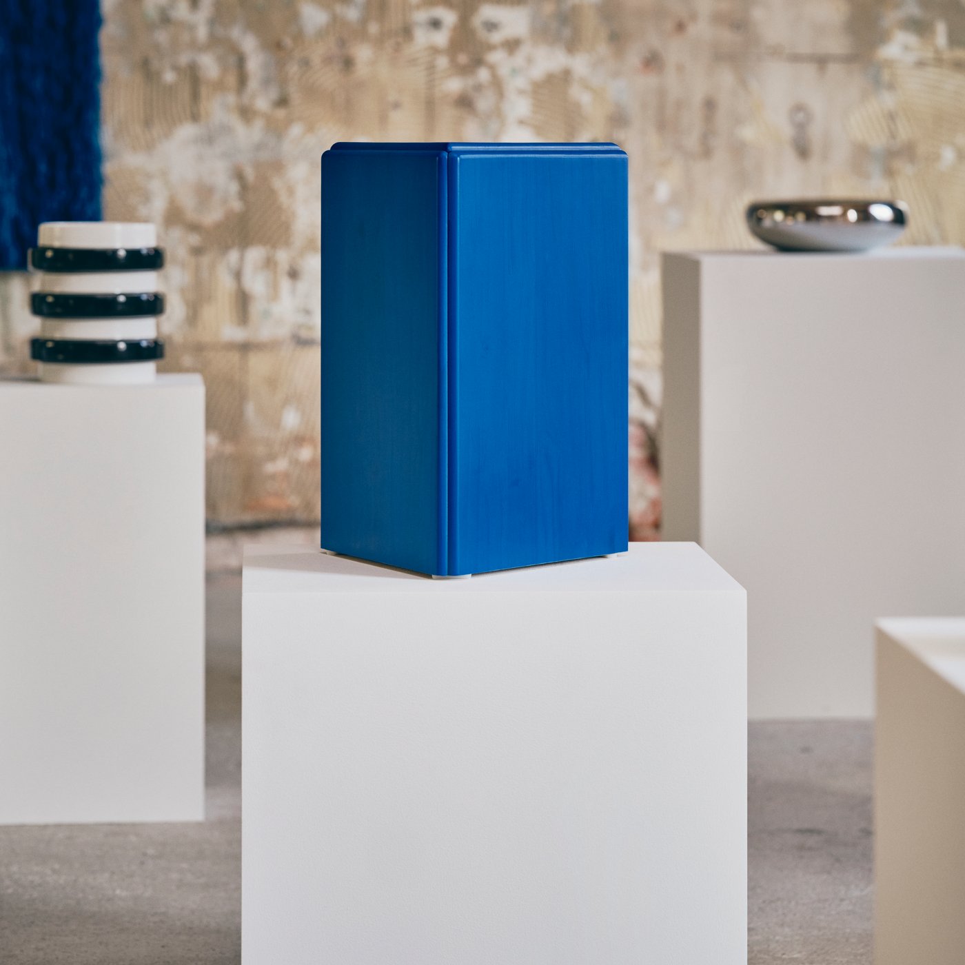 Wooden block painted blue is displayed on a white plinth in an industrial gallery, with additional black and white sculpture and chrome silver bowl in the background.
