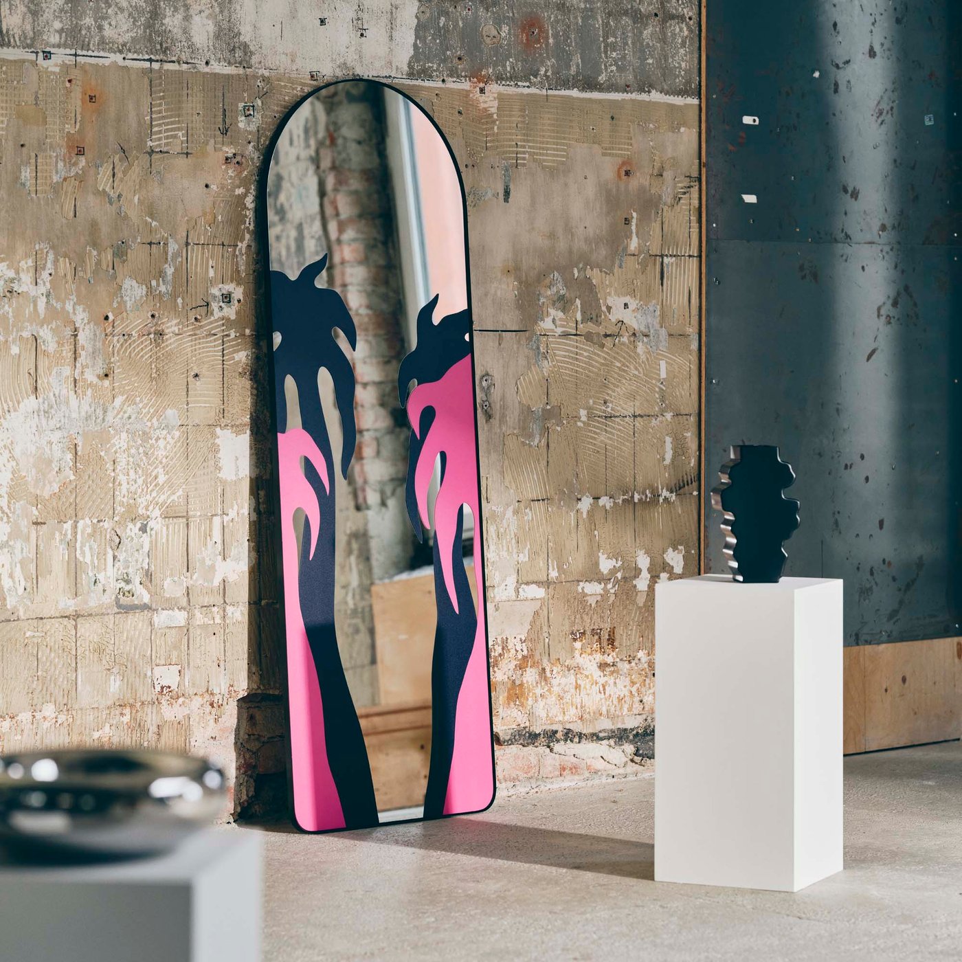 A tall, arched mirror with a foil print decoration stands propped against a brick wall, next to two objects on plinths.