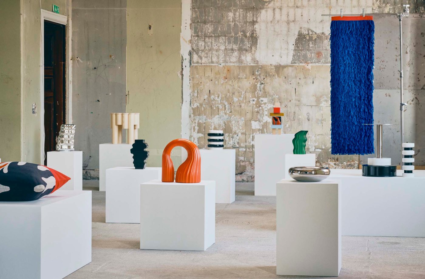 A landscape view of an industrial style gallery setting filled with colourful objects positioned on plinths.