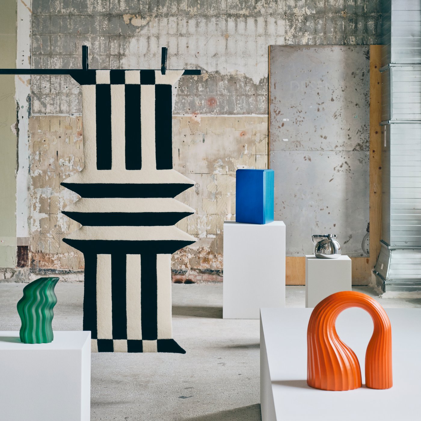 Contemporary art exhibition featuring a large black-and-white geometric rug hung from the ceiling, colorful abstract sculptures, and white display plinths in an industrial gallery.