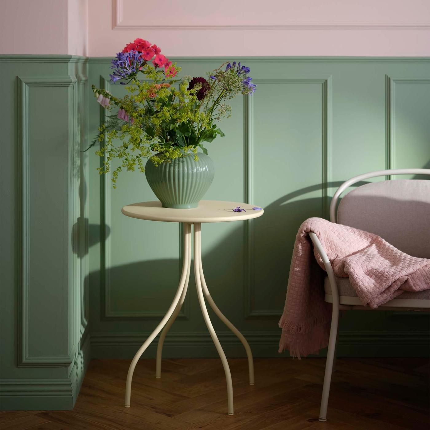 A vase of flowers on a light grey / beige side table in front of a green and pink wall.