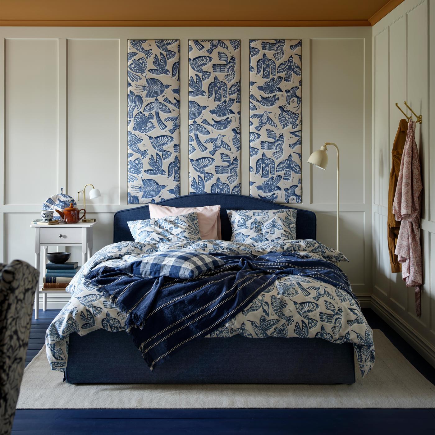 A cozy bedroom features a bed with blue and white patterned BITTERSÖTA bedding. A blue throw is draped over it. The headboard is upholstered in dark blue, complementing the decor. Panels of BITTERSÖTA fabric are hung above, and a nightstand is visible to the left.