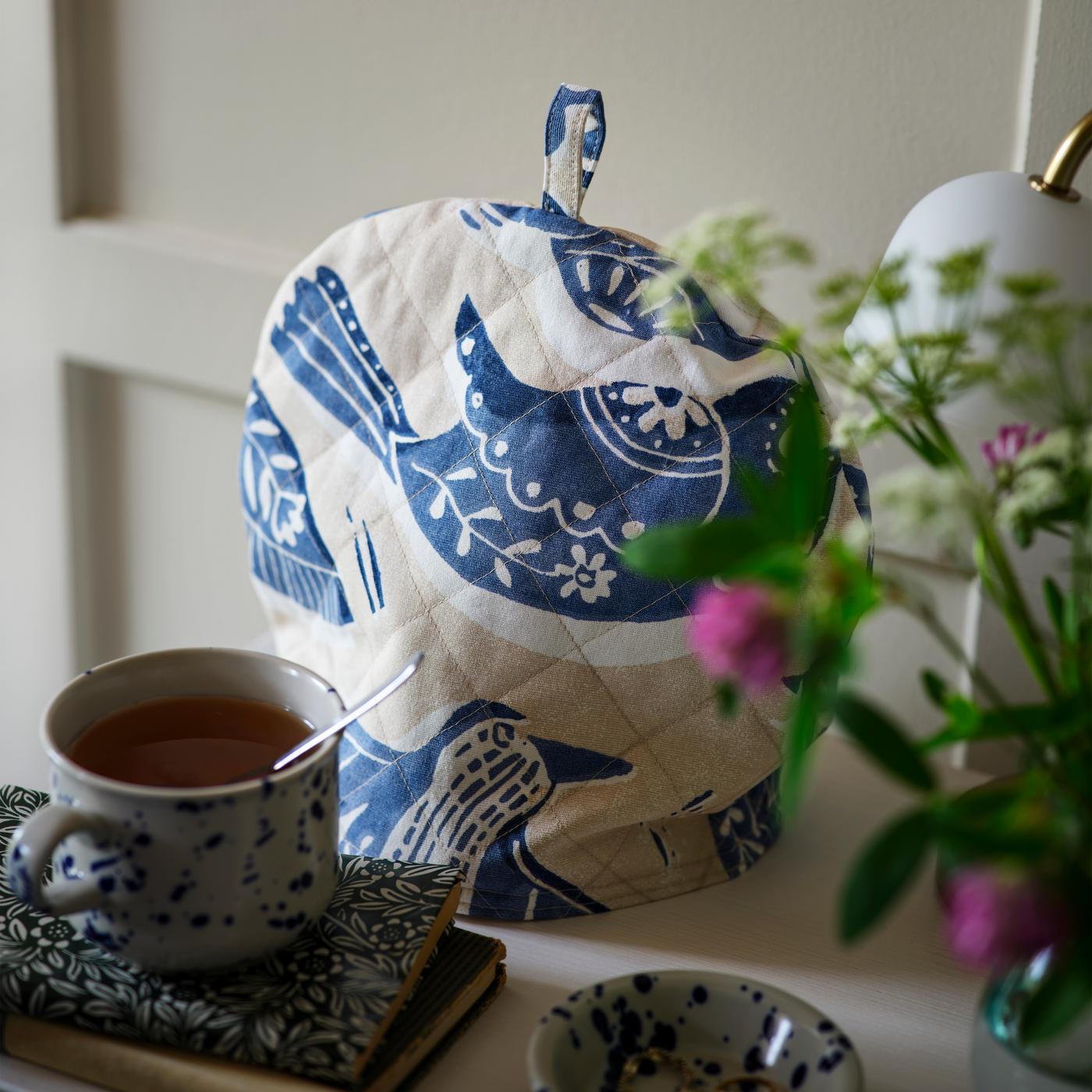 A tea set with a cup and saucer featuring a delicate pattern sits alongside a teapot covered with a cozy made from BITTERSÖTA fabric. Nearby, a vase holds fresh pink and white flowers, creating a serene and inviting tea-time scene.