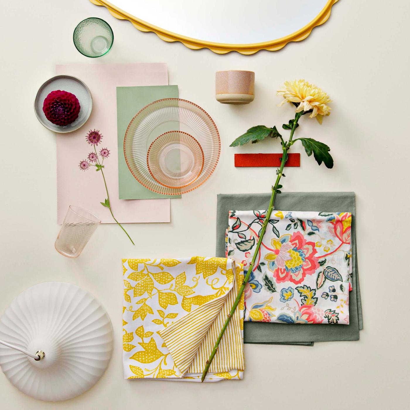 Moodboard flat lay featuring green, pink and yellow tones, with floral bedding, a fresh flower, glass bowls and a scalloped yellow mirror edge.