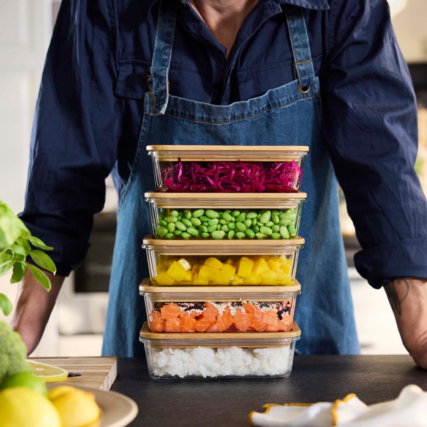 A stack of glass food containers with lids containing different foods on a surface. A person in an apron stands behind the containers.