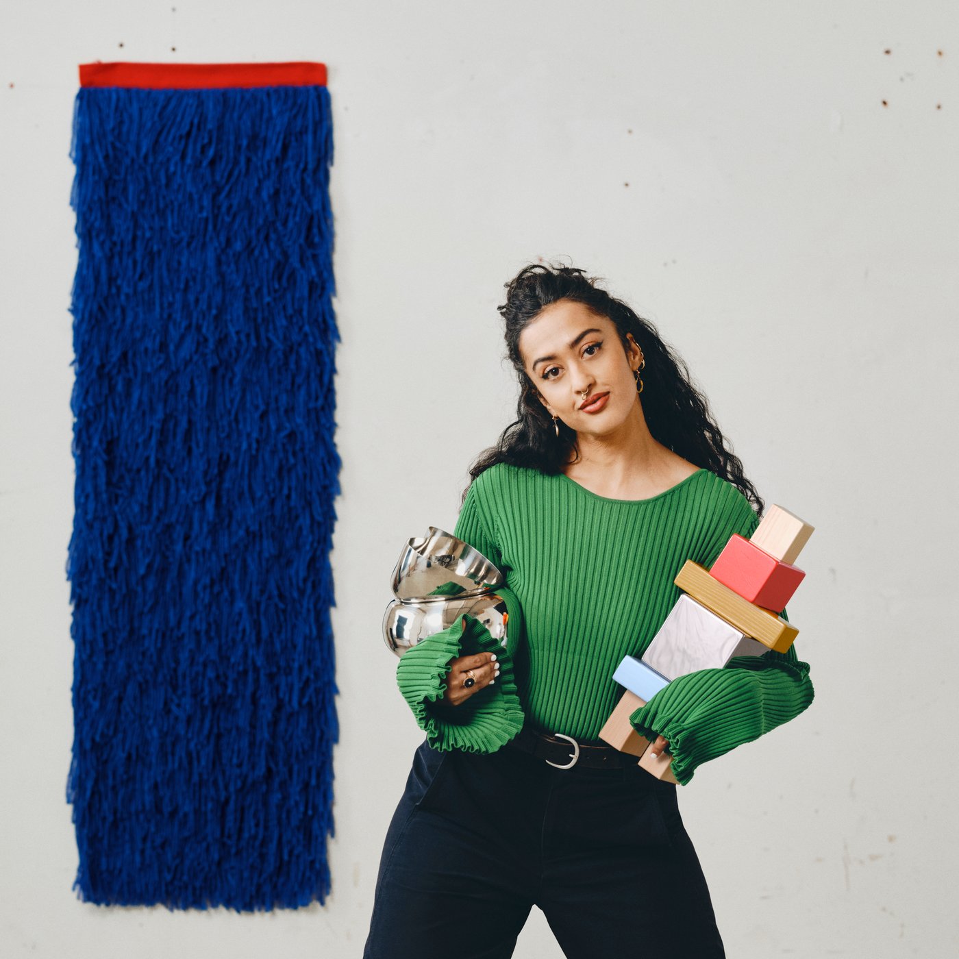 Designer Akanksha Deo holding three objects she designed for KONSTRUNDA including a wall hanging, a sculpture and a stainless steel bowl