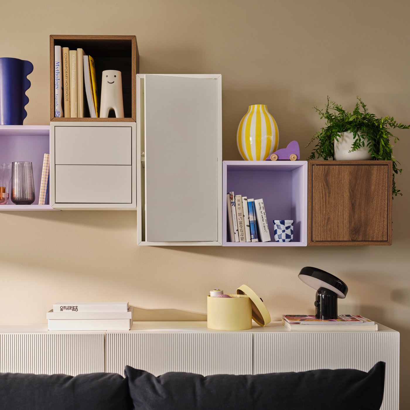 Open and closed EKET wall-mounted storage units in white, walnut effect and pale lilac on a beige living room wall.