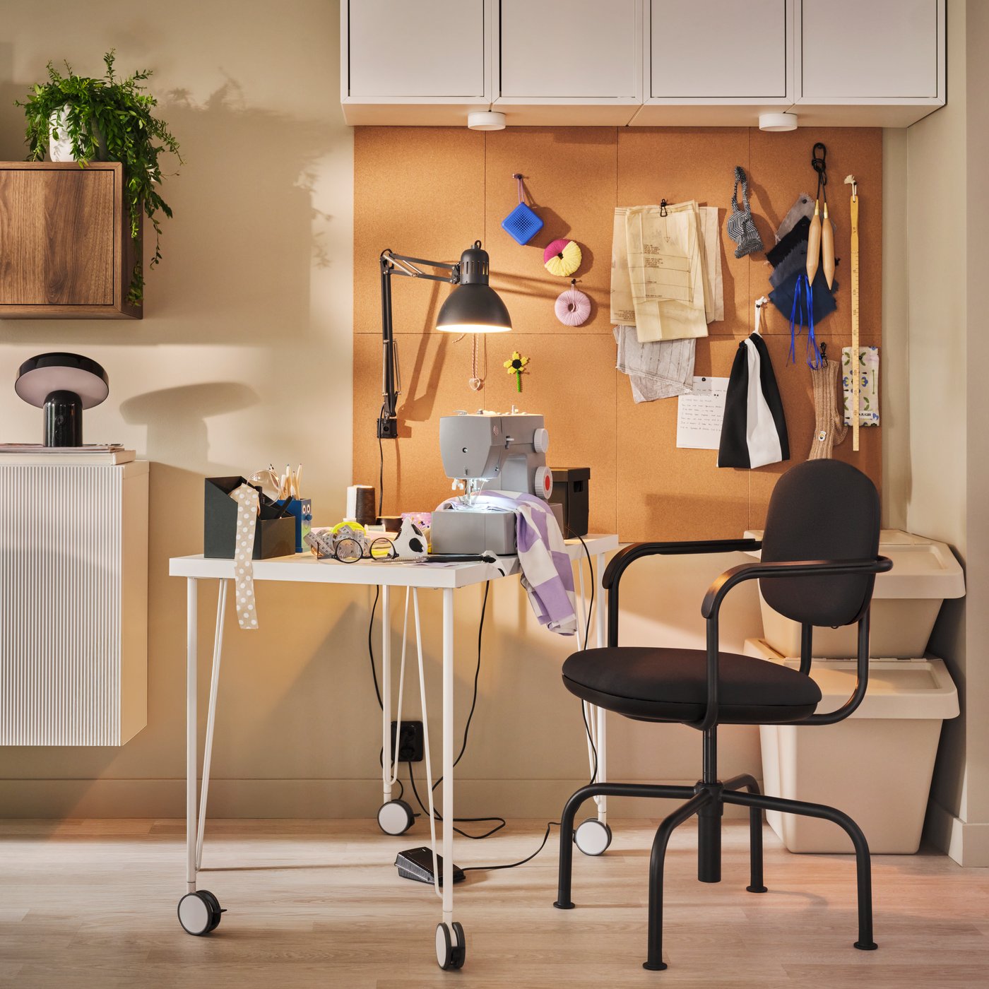 A sewing machine rests on a white desk beside a black office chair in front of a cork board in a creative workspace.