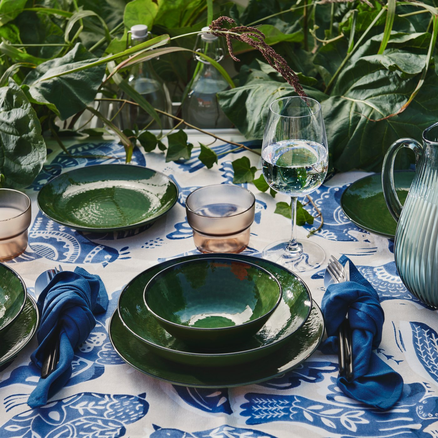 An outdoor table setting featuring green tableware on a blue and white decorated tablecloth, surrounded by greenery.