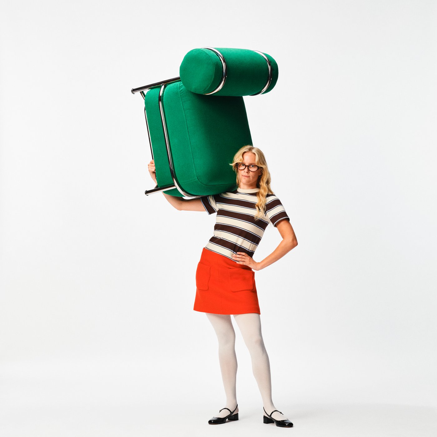 Person standing against a white background, carrying a green upholstered chair balanced on one shoulder.