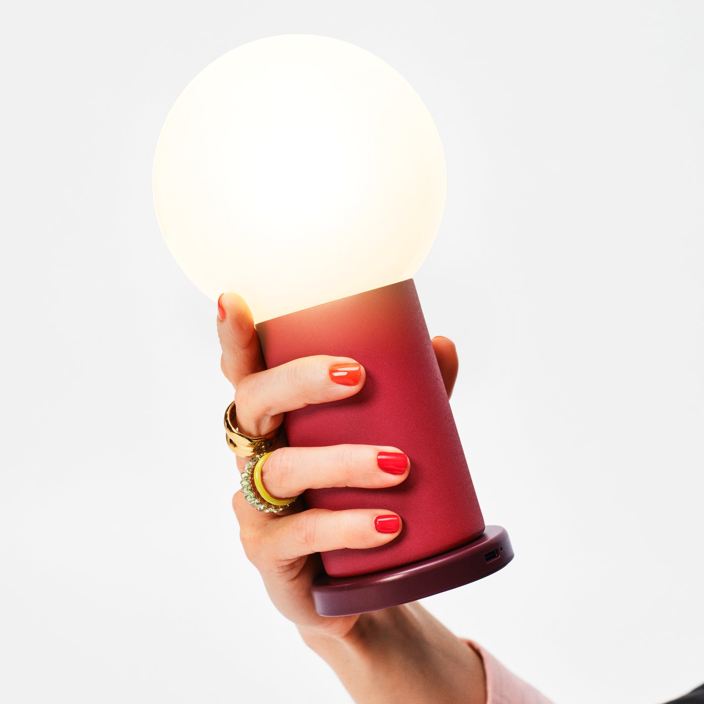 Hand holding a portable table lamp with a round glowing white shade and a matte red cylindrical base.