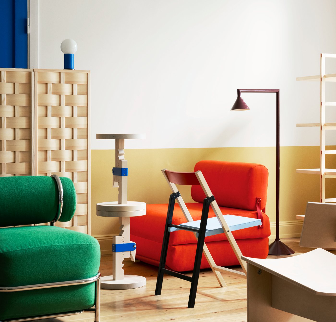 Interior scene with colorful furniture: green lounge chair, red armchair, floor lamp, shelving, and a modular wooden side table.