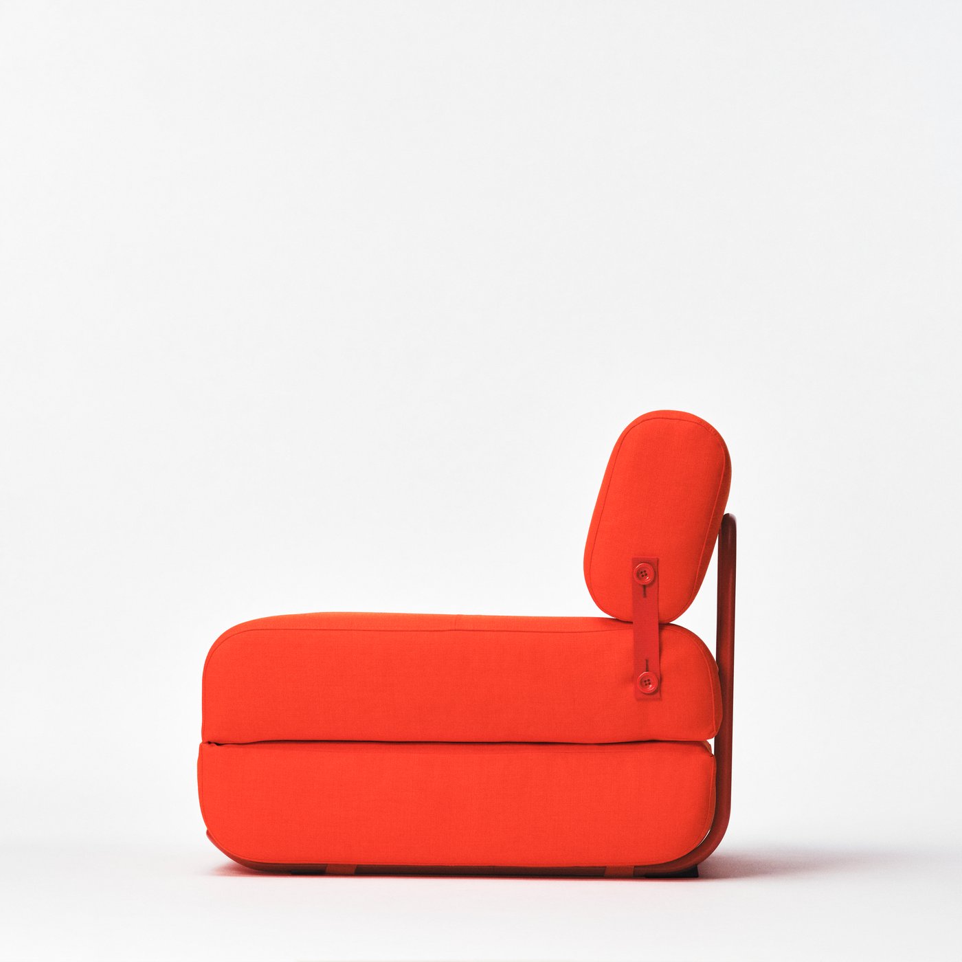 A red upholstered easy chair with a steel frame and strap is seen from the side.