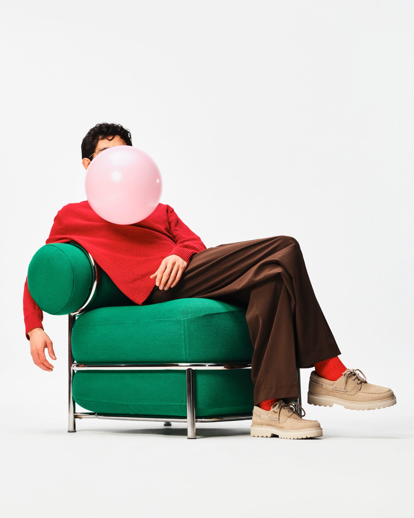 Person reclining in a green upholstered chair, blowing a pink bubblegum bubble, against a white background.