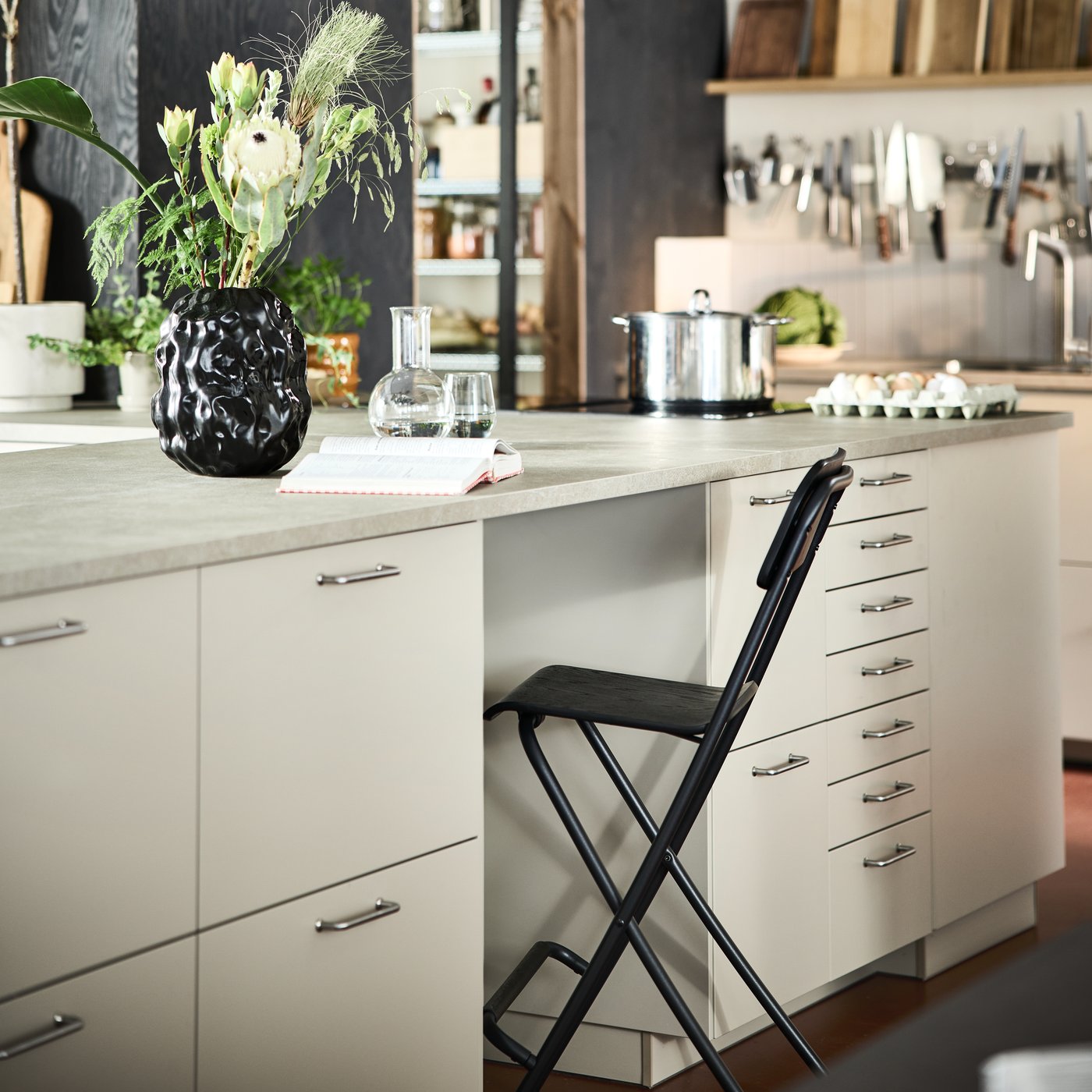 A white ASPUDDEN kitchen with a close-up of a kitchen island with lots of drawers, a grey countertop with an open space for a black folding chair, with some decor and organised kitchen accessories in the background.
