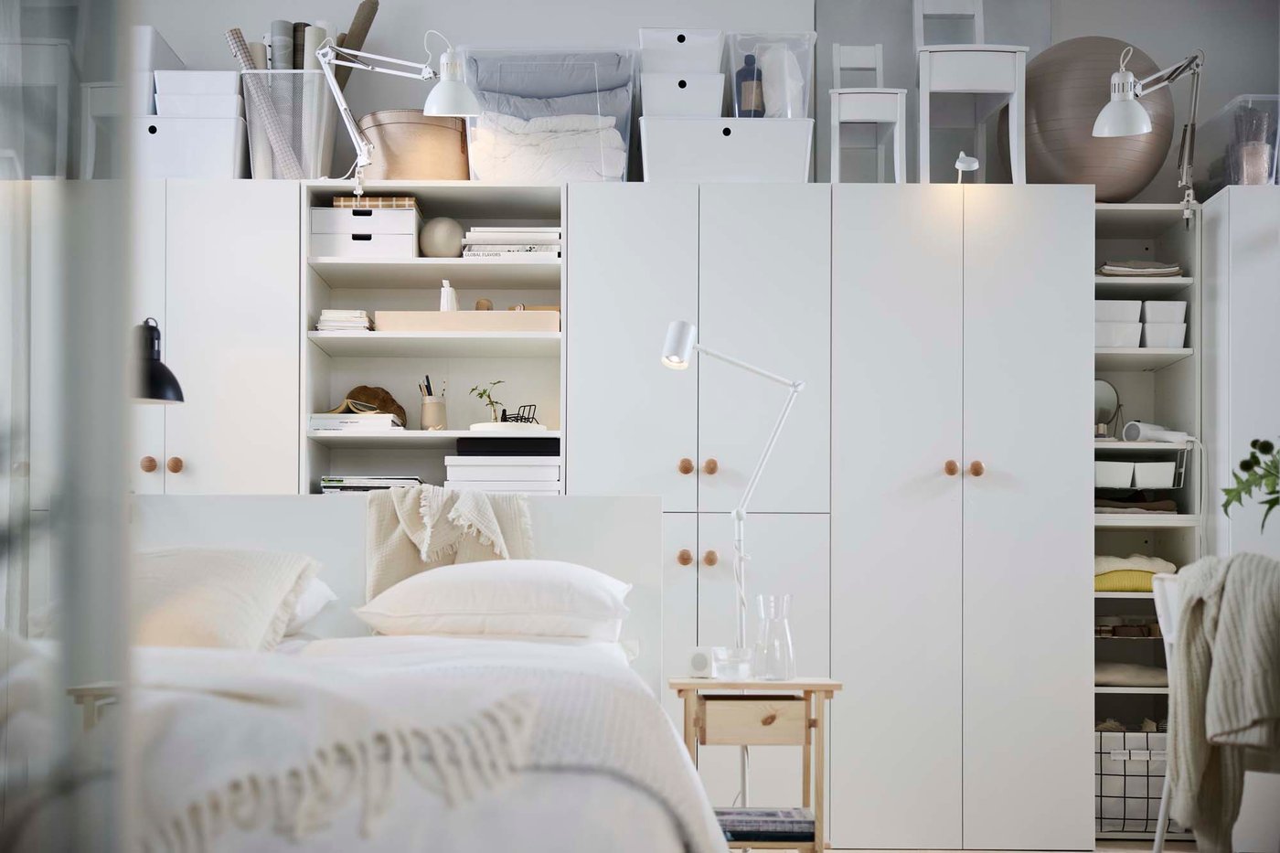 A white studio with a bed in the foreground and a full-height storage solution along the length of the back well.