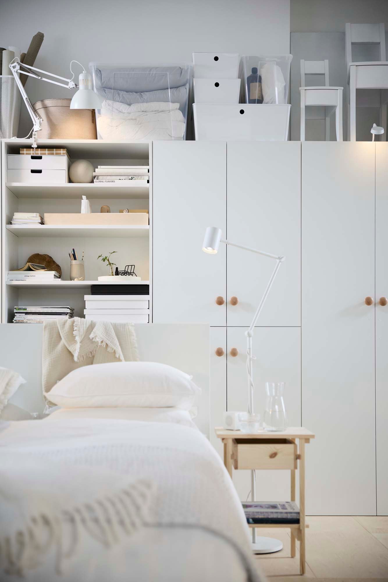 A wooden bedside table beside a white bed in front of open and closed white wall-to-wall storage.