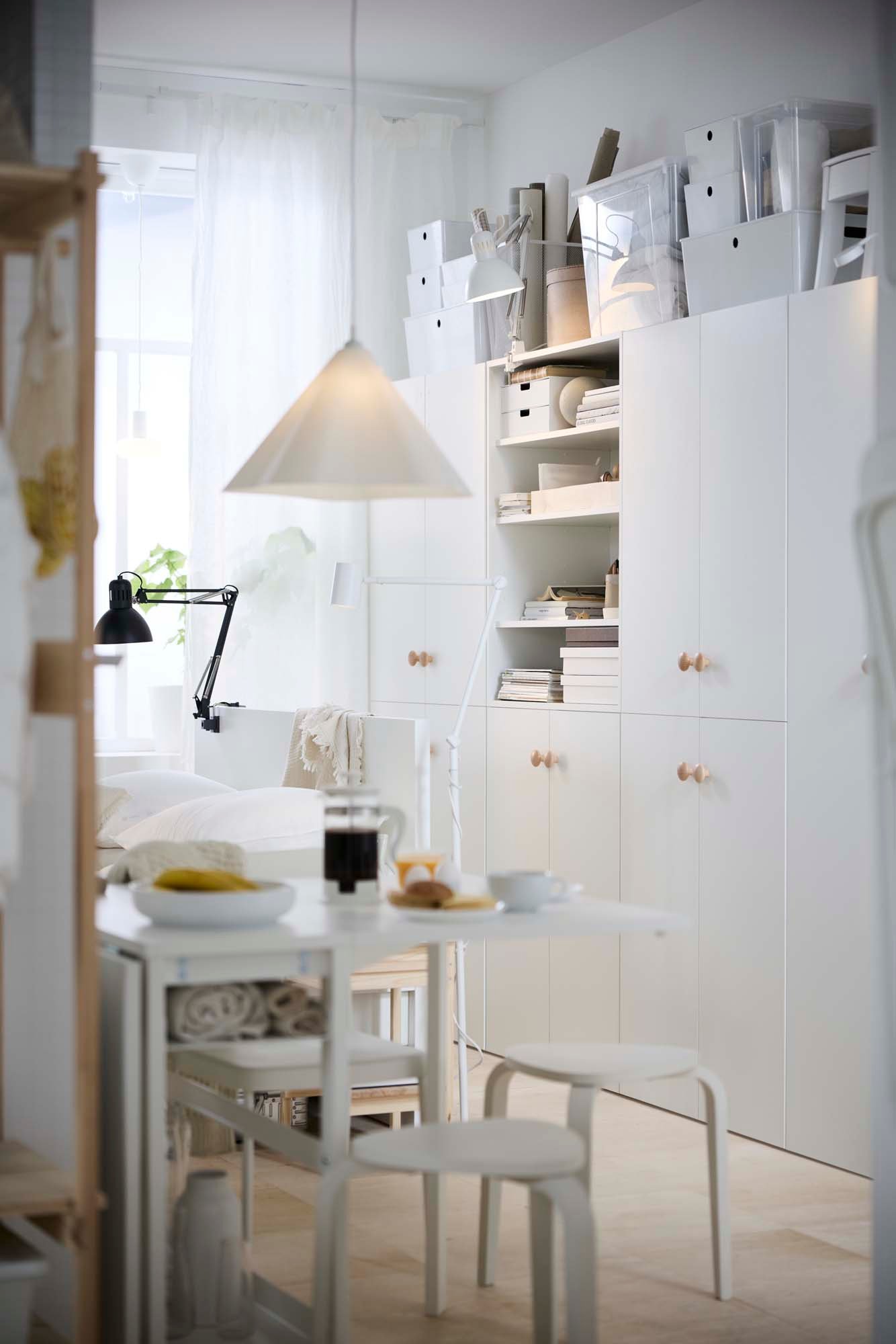 A white gateleg table surrounded by white stools in front of a white storage system.