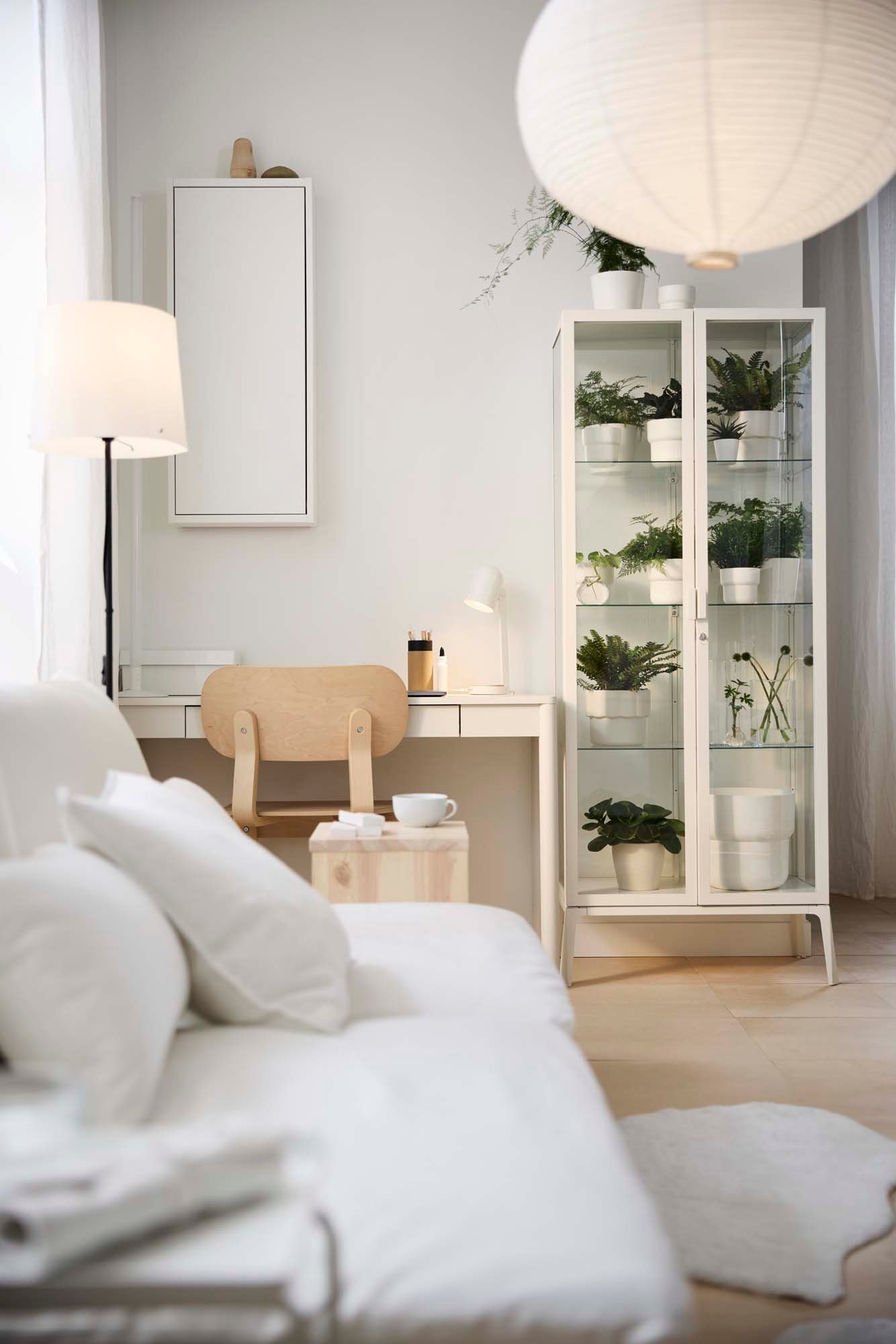 A white desk with a closed white wall mounted cabinet above and a white glass-door cabinet beside.