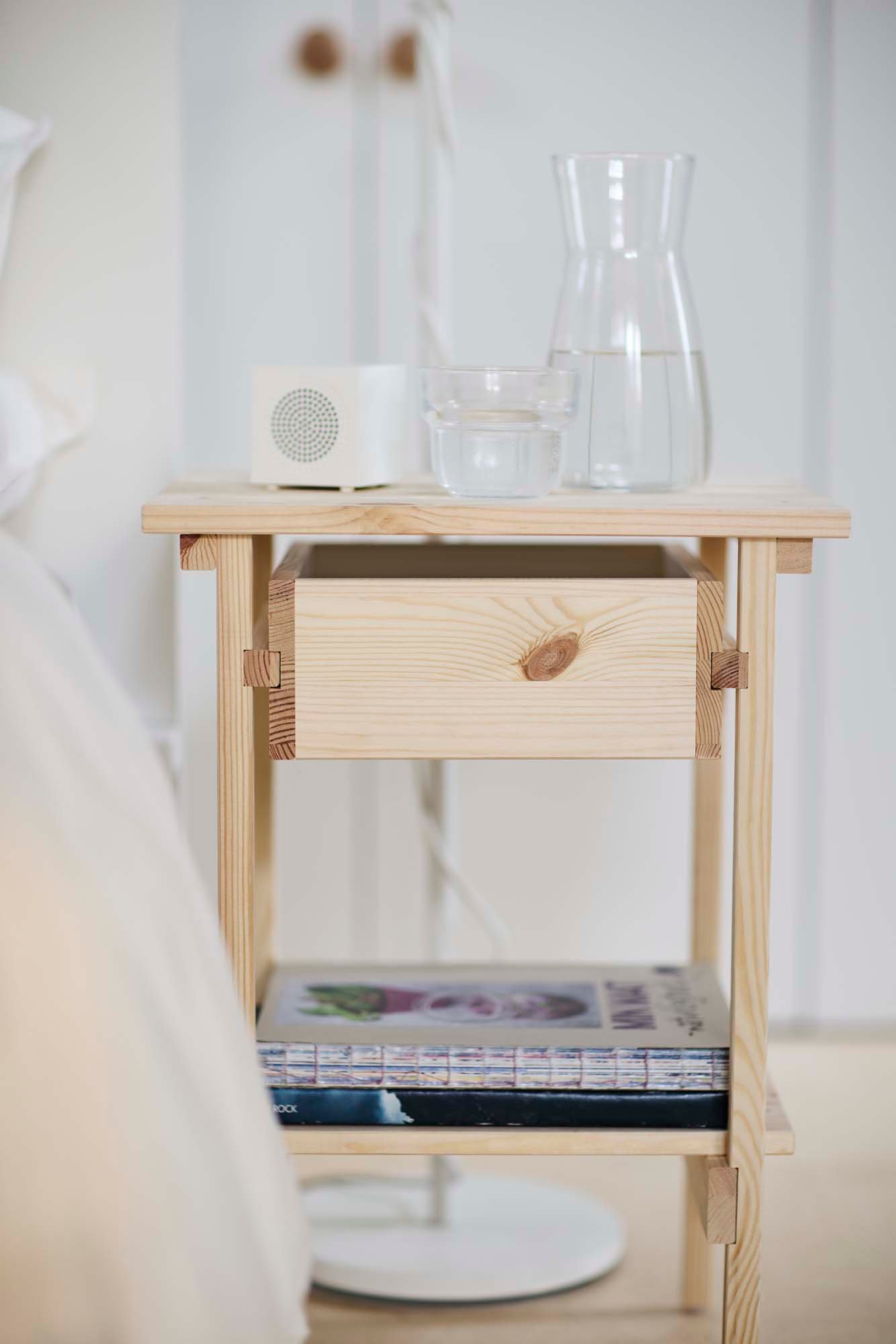 A speaker and vase with water on a light wood bedside table,