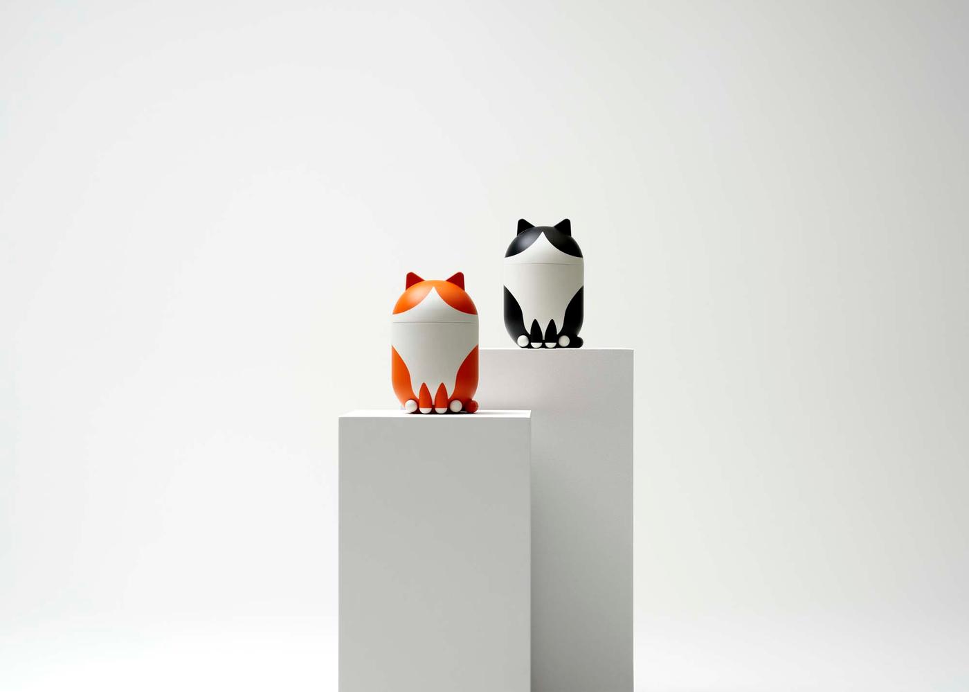 Two cat-shaped storage containers with their lids on plinths in a white environment.