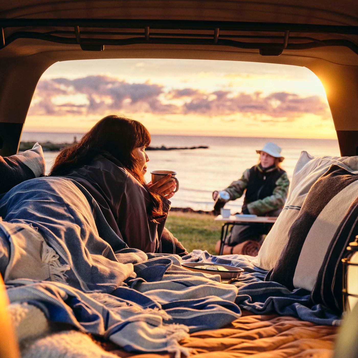 A person lying under a throw sips from a mug in the boot of a car while looking out to sea.